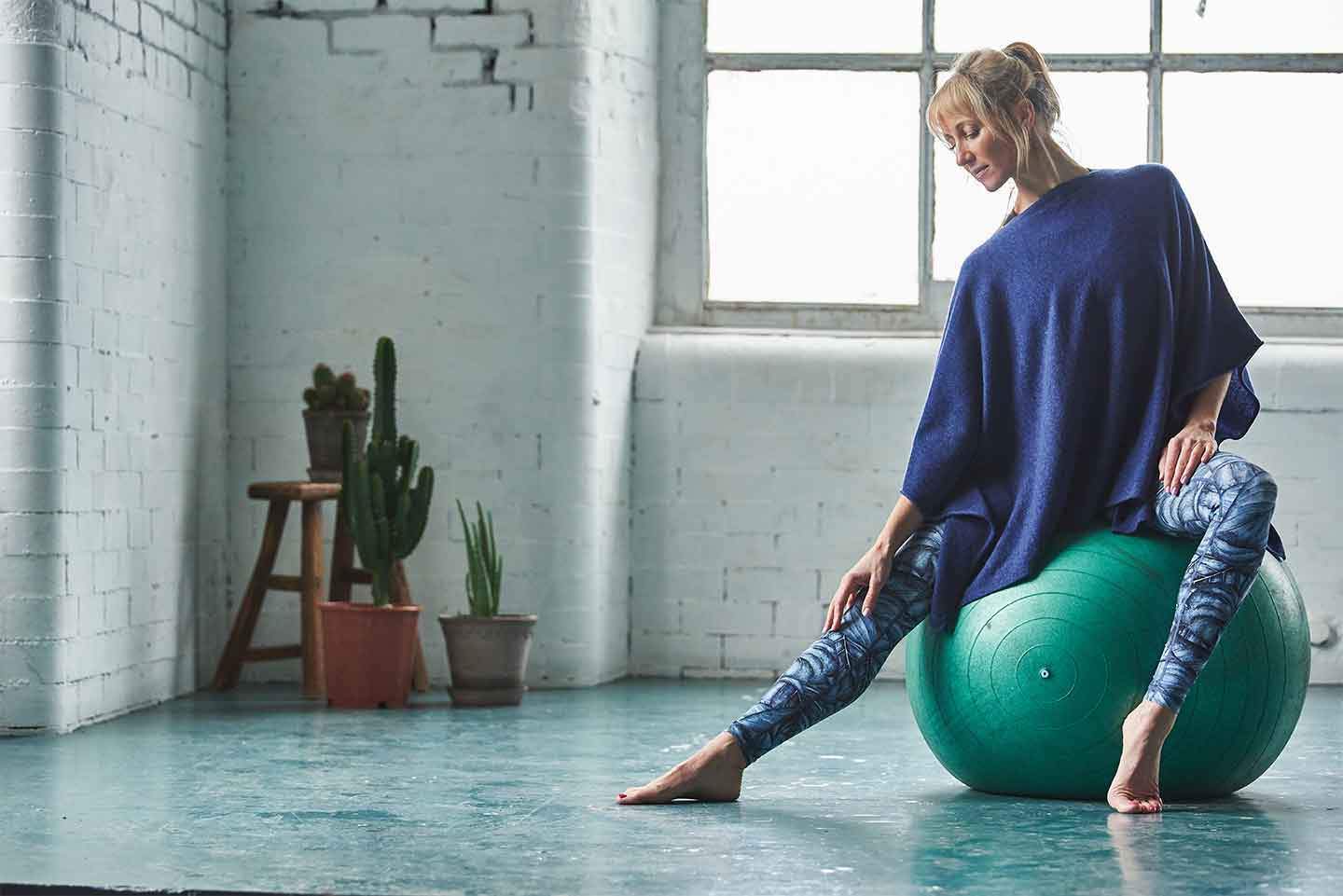 Women-doing-yoga-in-front-of-a-window-sat-on-a-yoga-ball-in-sportswear-and-blue-cashmere-poncho
