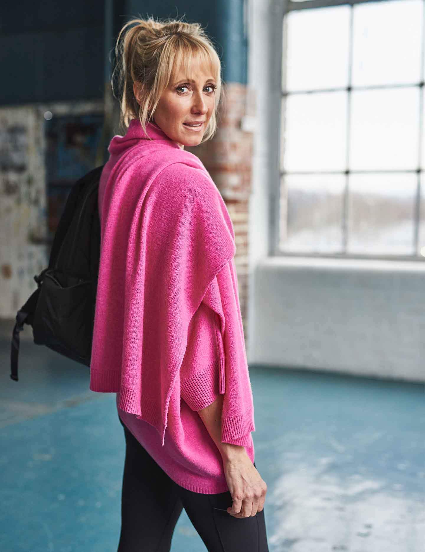 Women standing in pink cashmere jumper with pink cashmere scarf wrapped around shoulders with black rucksack on back