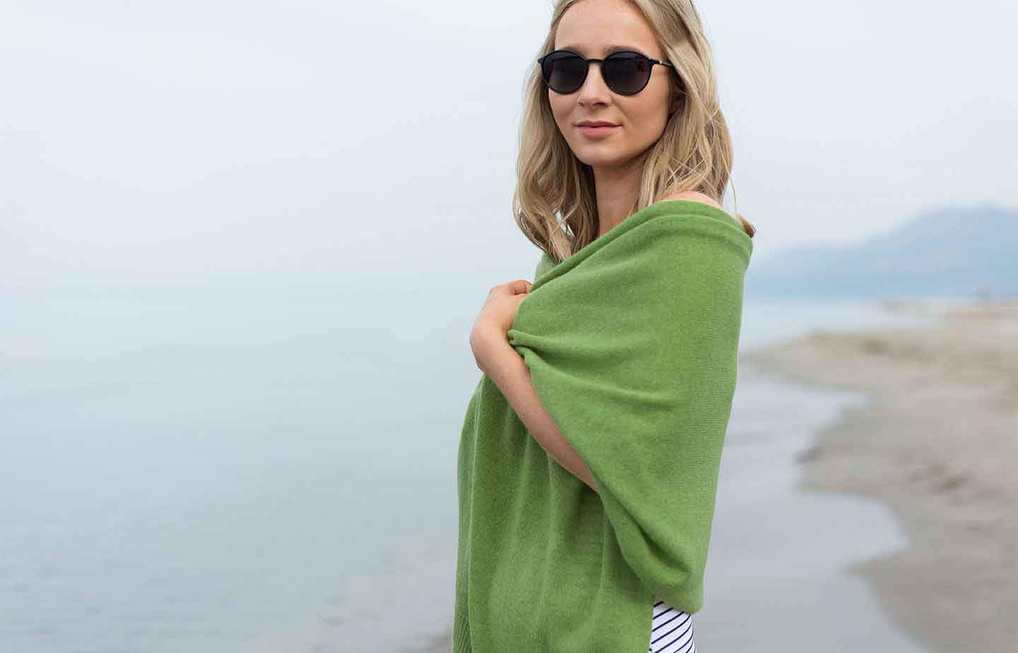 Women-standing-on-a-beach-with-blue-sky-in-the-background-wearing-sunglasses-and-clutching-a-green-cashmere-wrap-around-her-shoulders