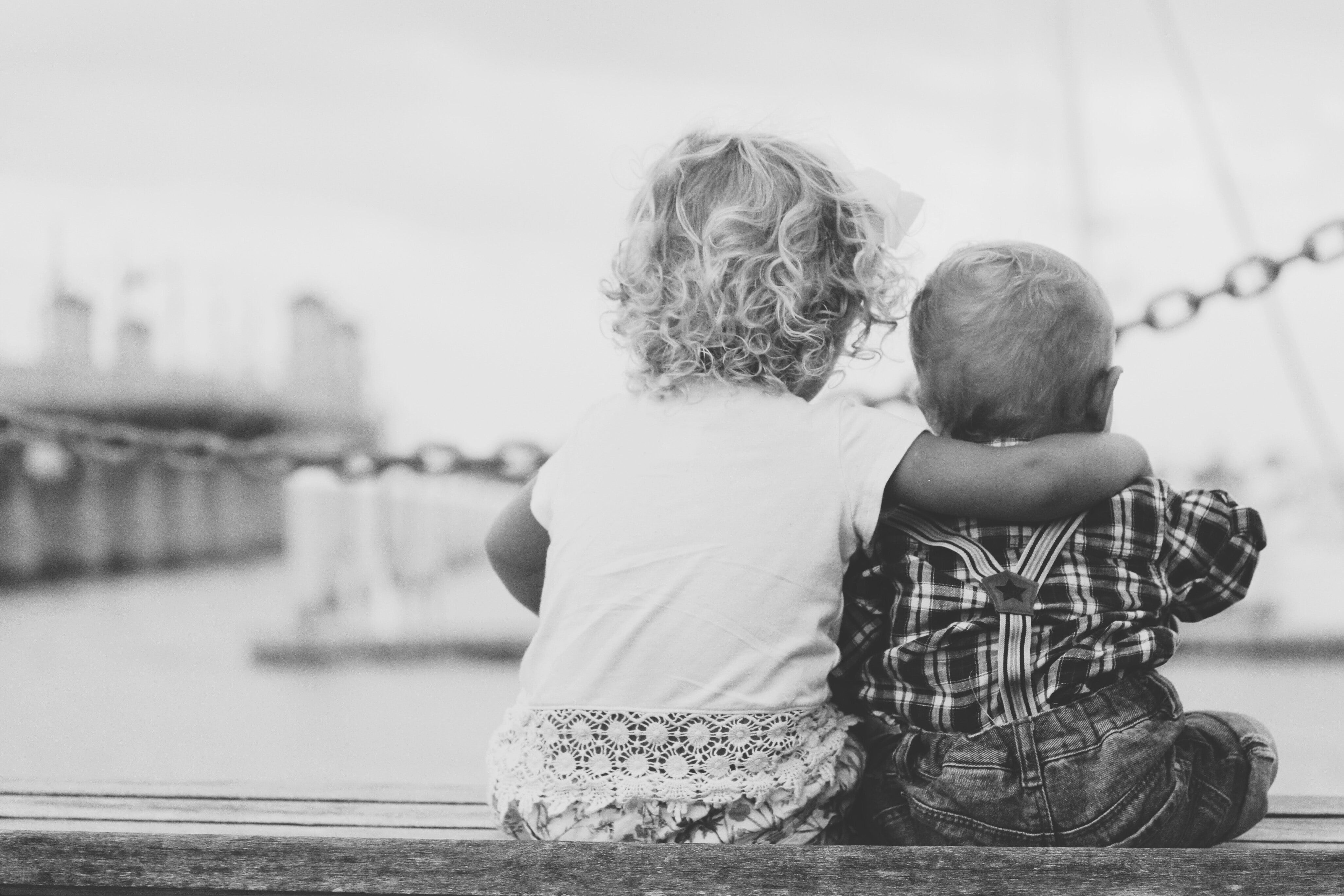 Black & White Photo of Toddlers Arm-In-Arm Facing Away From Camera