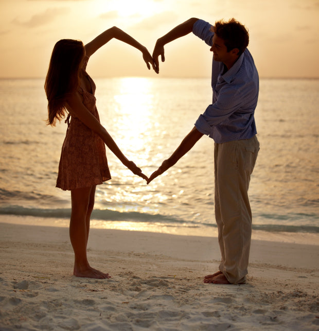 Man and woman on the beach at sunset making a heart with their joined arms