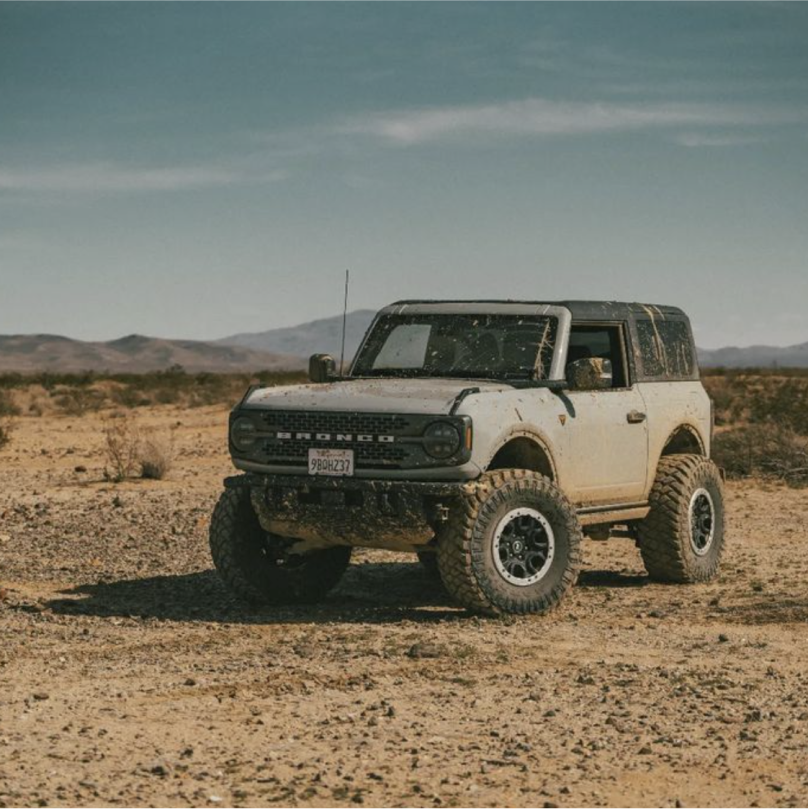 A silver and black Ford Bronco parked in the middle of a sandy desert covered in mud and sand from offroading.