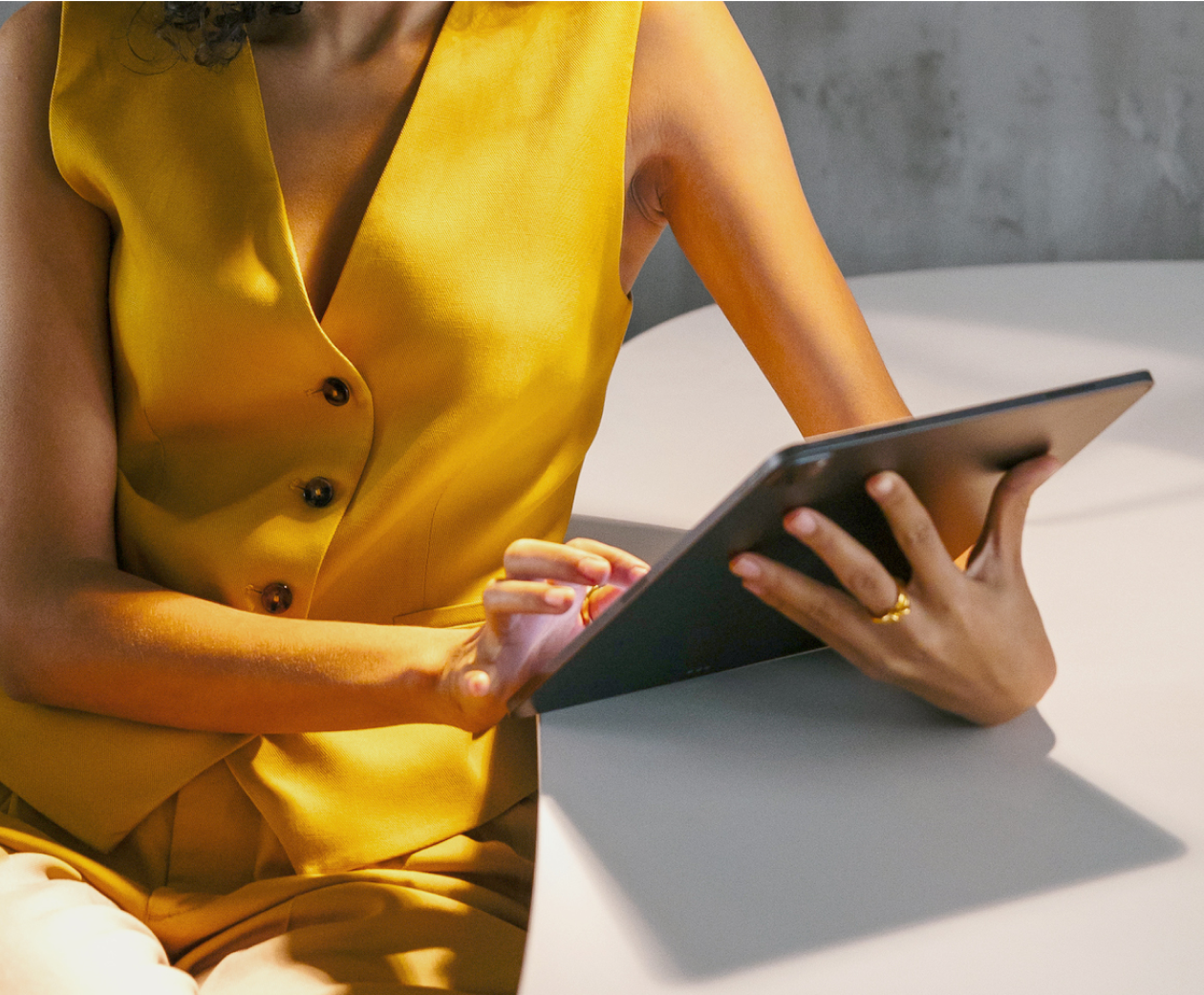 Woman with medium complexion, shown from the neck down, sitting at a table and working on a tablet..