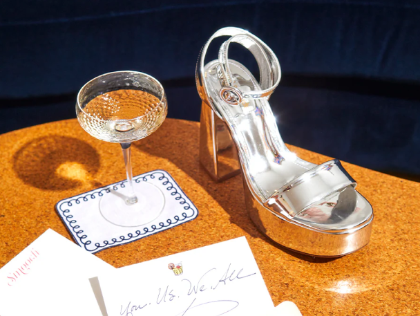 A stylish Larroudé silver high-heeled sandal placed on a table next to a filled champagne glass on a coaster, with a handwritten note and an envelope nearby.