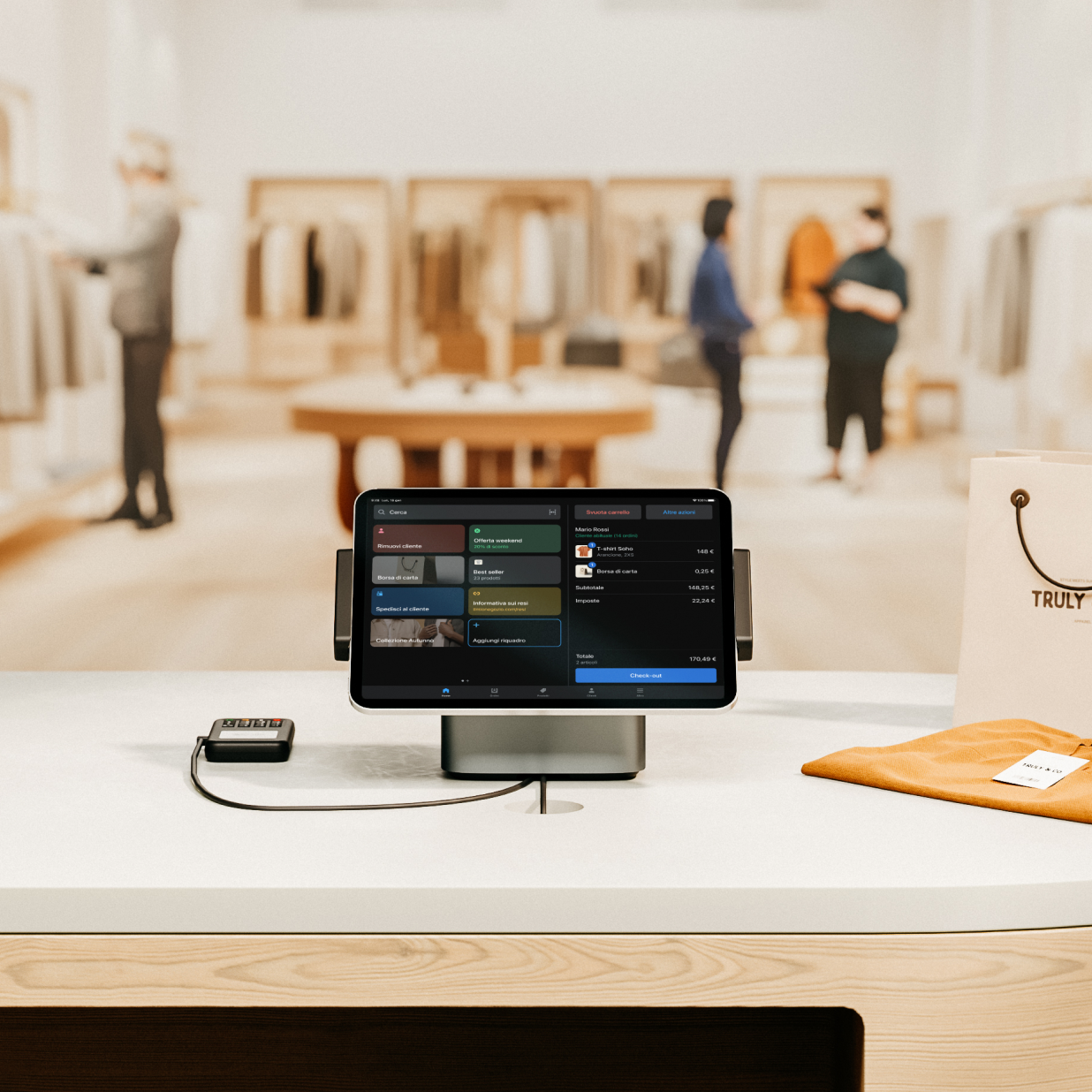 Veduta da dietro al bancone di un grande negozio di abbigliamento chiamato Truly and Co, in un magnifico centro commerciale illuminato. Sul bancone ci sono due configurazioni di Shopify POS che mostrano l’app POS su un tablet, che è connesso direttamente a POS Terminal. Nel negozio ci sono persone che fanno acquisti e un commesso che aiuta un cliente a effettuare il check-out in negozio.