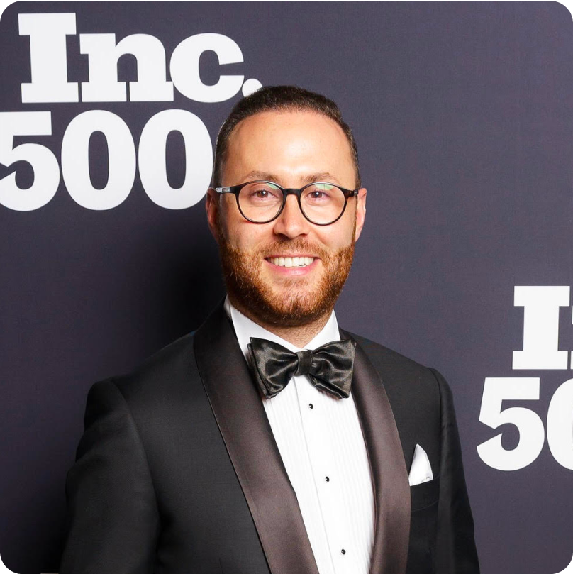 Hatching Time owner Yagiz Aksu, dressed in a black tuxedo and bowtie, smiles in a photo op in front of a backdrop at an Inc. 5000 gala.