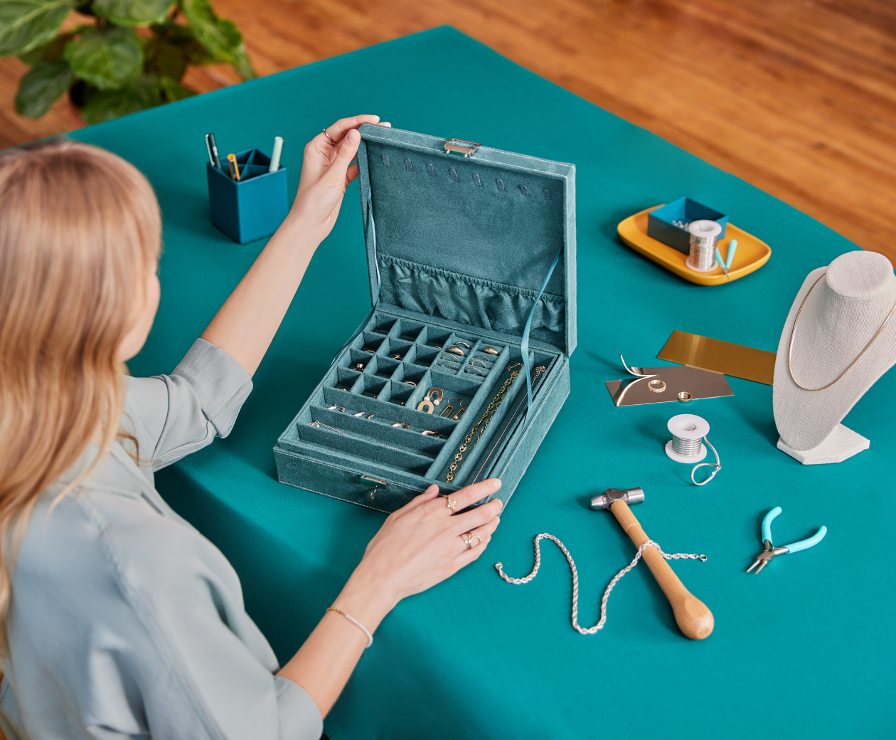 A white female Shopify merchant of a fine jewelry shop looking into a velvety teal box of necklaces, earrings, and rings, and repairing a few of the necklaces with her tools.
