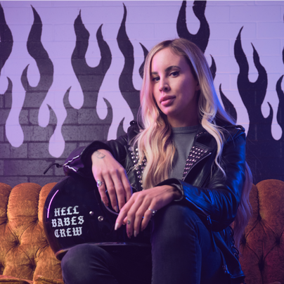 Jessica Wise, PDG de Hell Babes, assise sur un canapé vintage avec un casque de moto portant l’inscription « Hell Babes Crew » devant un mur de briques orné de flammes noires et violettes.