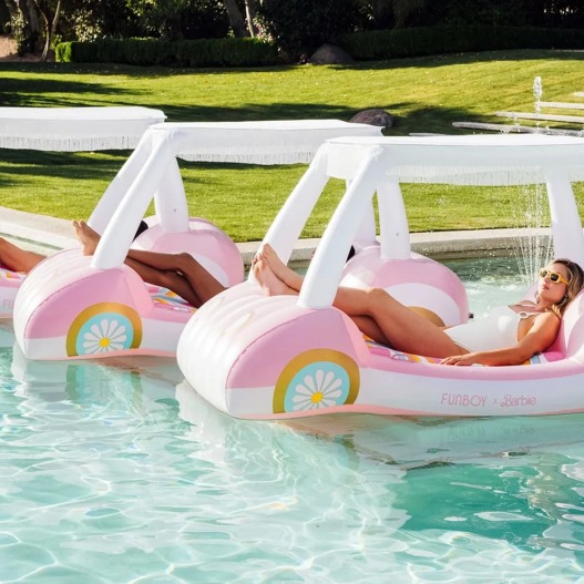 Two women relaxing on pink and white car-shaped pool floats from the Mattel and Funboy Barbie movie collaboration, floating in a clear pool with a grassy lawn in the background.