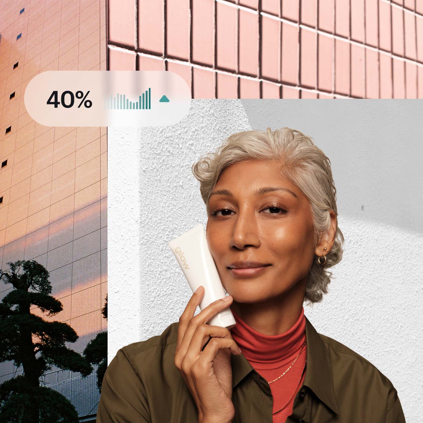 Photo collage. In the foreground, a close up of a merchant from the shoulders up, smiling in a relaxed and confident way, looking into the camera and holding a small white tube of skincare product next to their cheek. A small graphic shows a chart with upward trajectory next to 40%. The background photo features a tall window-covered building reflecting a pink sunset, with one dark tree in the foreground.