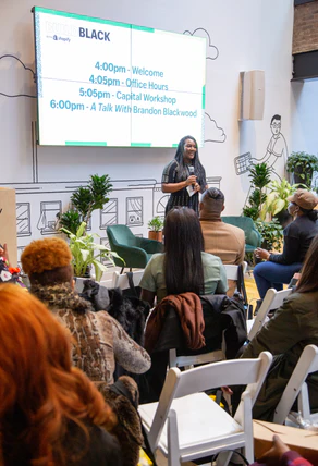 A photo of the Build Black program lead, holding a mic infront of an audience of seated guests at a Build Black event. There's a large monitor behind the program lead indicating the schedule for the event.