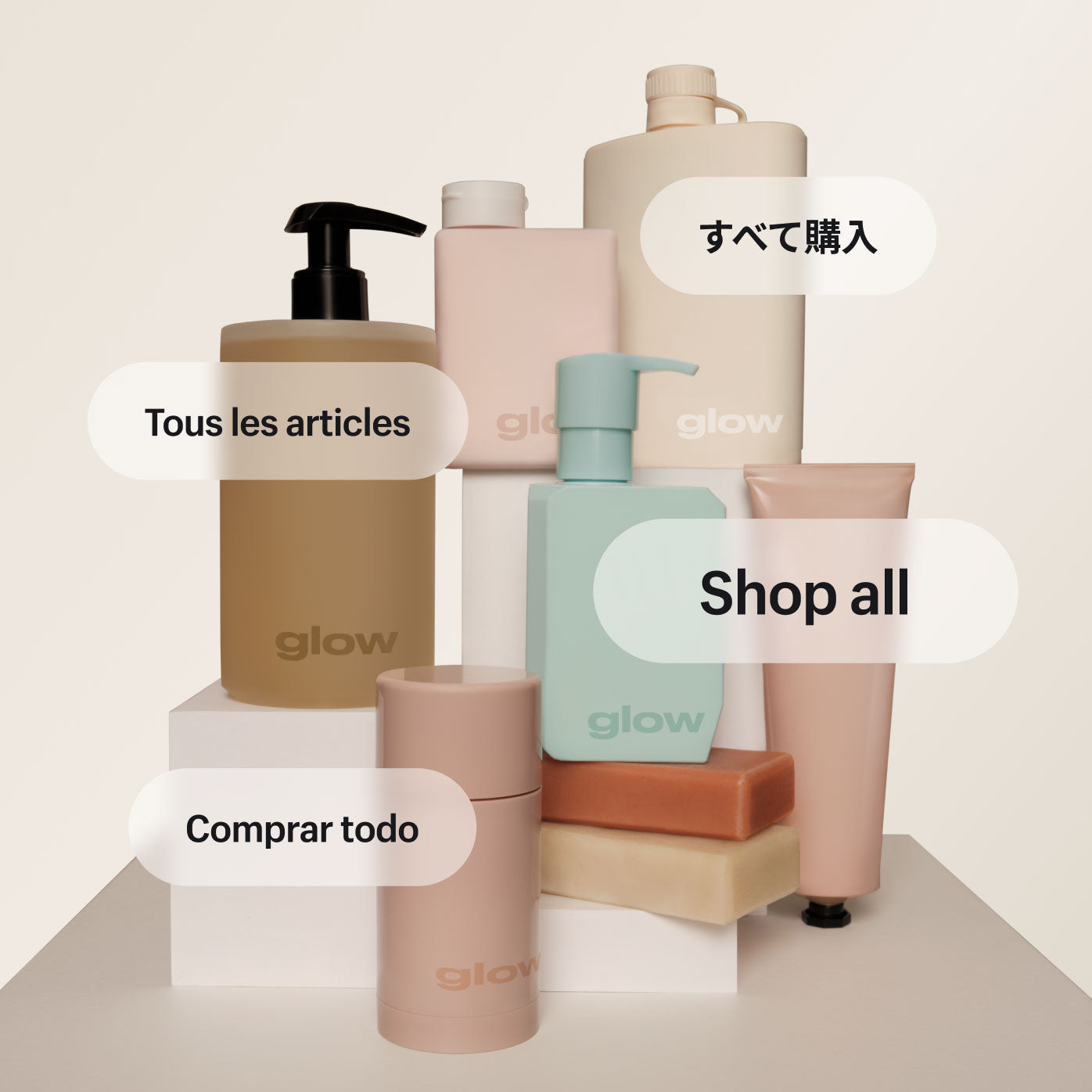 A collection of skincare products in stylish neutral-toned minimal packaging assembled on a platform. Products range from bottles and tubes to bars of soap and a cylindrical stick of deodorant. Each product is branded with a Glow logo. Several text graphics show the words Shop all translated into Spanish, French, and Japanese.