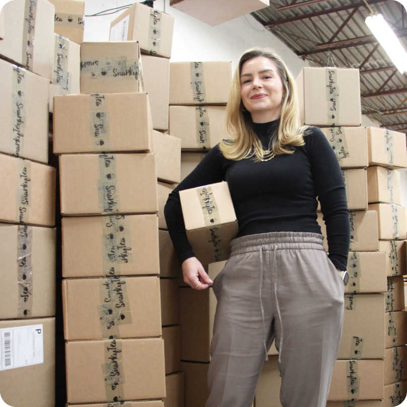 Jenni-Lyn Williams, CEO de SnarkyTea, sosteniendo una caja de envío, cerca de cajas de cartón apiladas