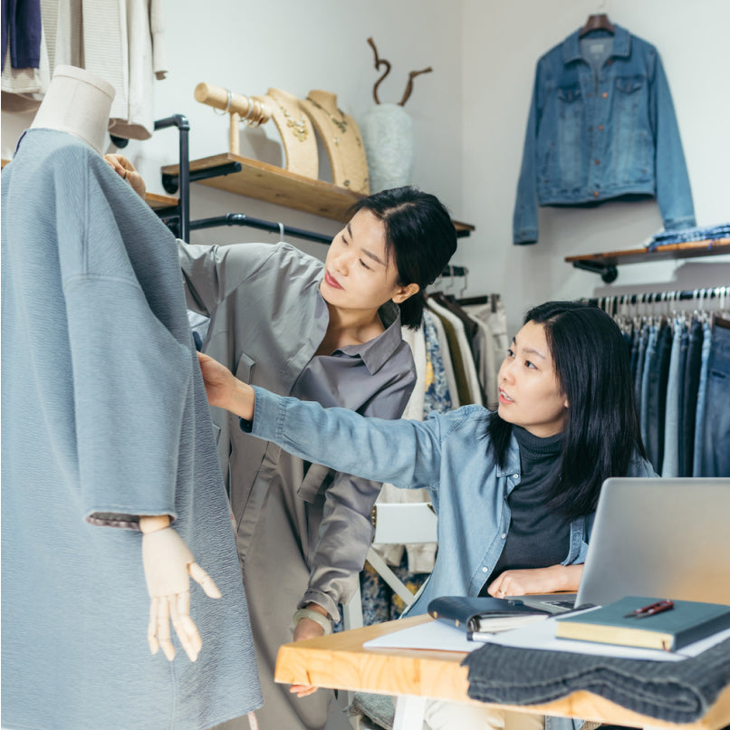 Twee vrouwen in een winkel die een jas op een paspop aanraken, met spijkerbroeken en jassen op de achtergrond.