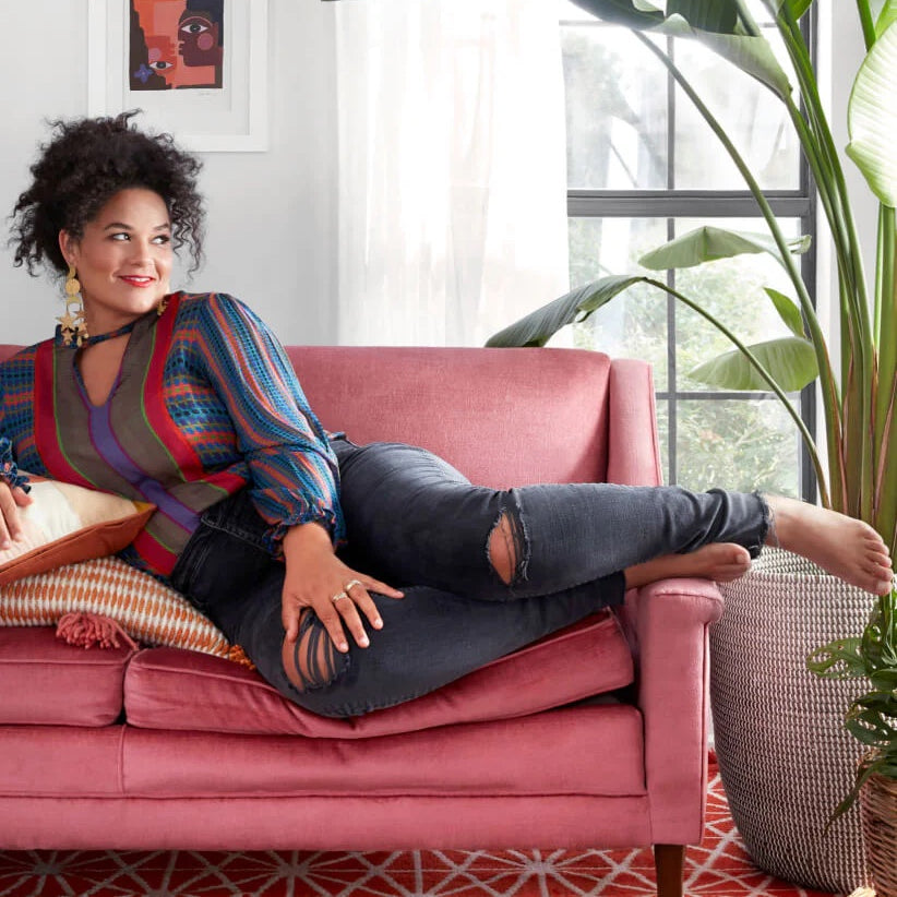 Founder of The Jungalow, Justina Blakeney, sits in a tall wooden chair in a room nicely decorated with many colourful pillows and an assortment of decorative potted plants.