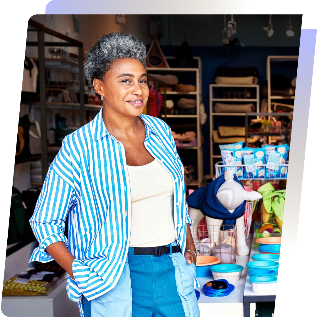 A confident woman with gray hair stands in her retail store, wearing a blue and white striped shirt, showcasing colorful products. This image represents the potential for growth with Shopify Capital.