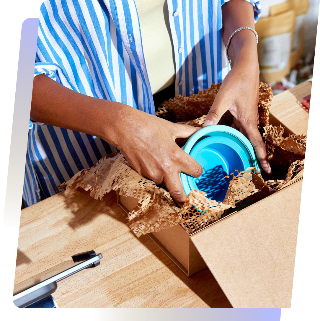 A close-up of hands unpacking a blue bowl from a cardboard box filled with packing material, showcasing the process of preparing products for customers in a retail setting, supported by Shopify Capital.