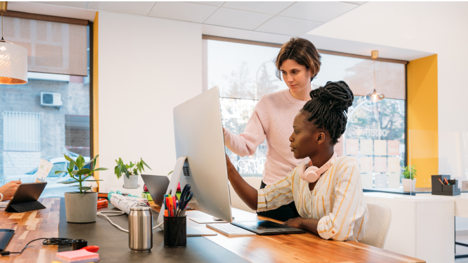 A white female and Black female Shopify merchant team reviewing their company budget on a large desktop in their retail office