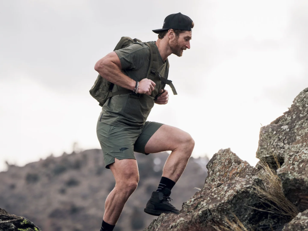 A man wearing Ten Thousand X GORUCK athletic gear, including a green t-shirt, shorts, and a backpack, climbing a rocky terrain in an outdoor setting.