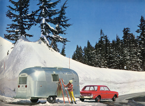 Airstream and red car in snowy setting, person standing nearby with skis