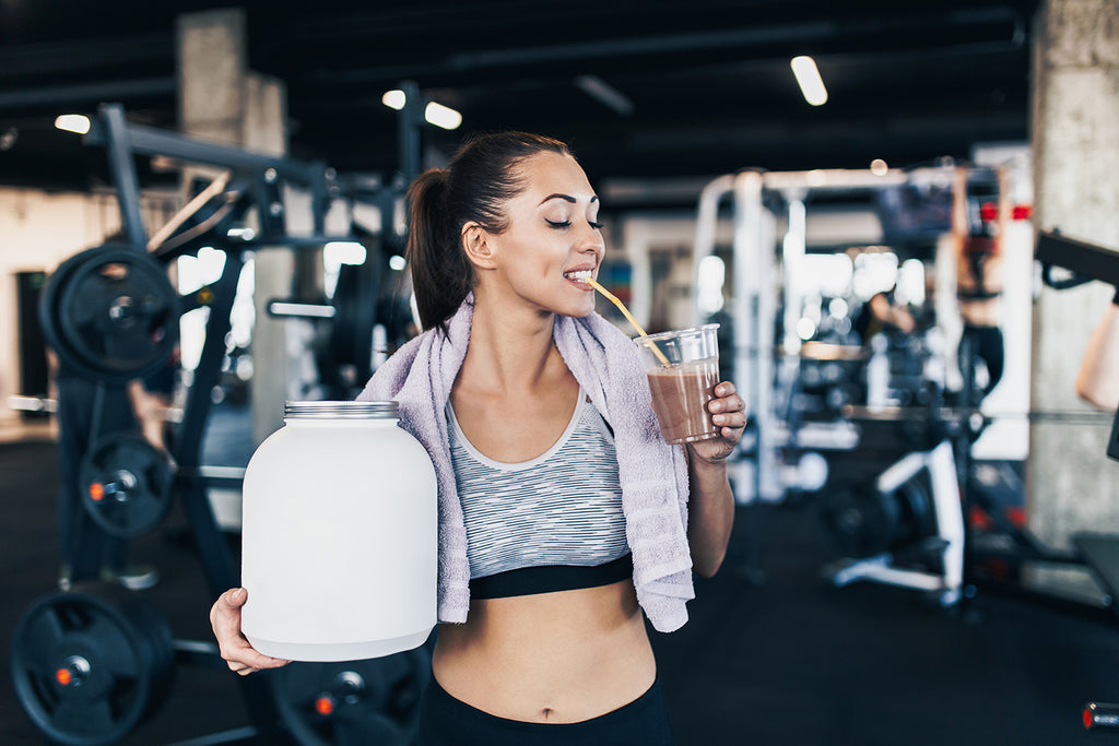 woman holding big white protein jar and drinks shake with drinking straw in other hand