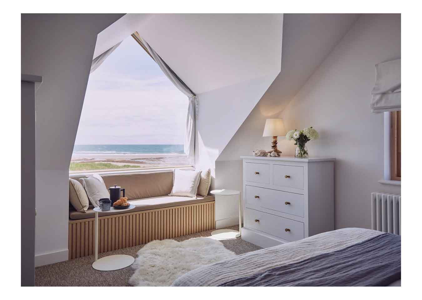 Bedroom with beige cushioned seating and oak wood cladding on the bay window, white furniture and a large window with a seascape outside.