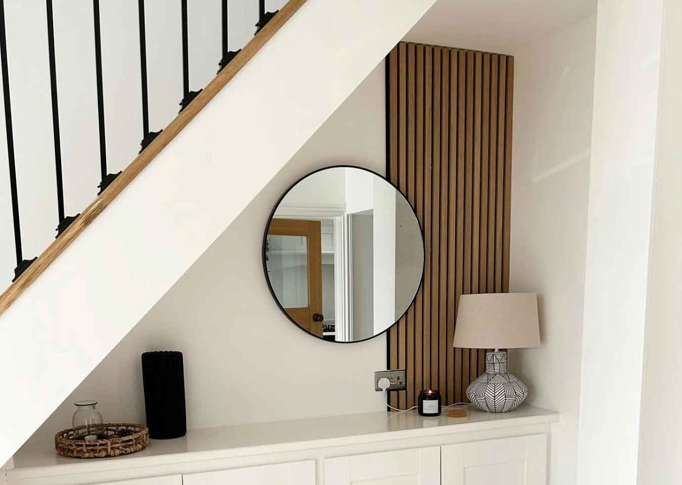 White under stairs storage featuring a single wood wall panel, a round mirror, beige lamp, black vase and a brown tray.