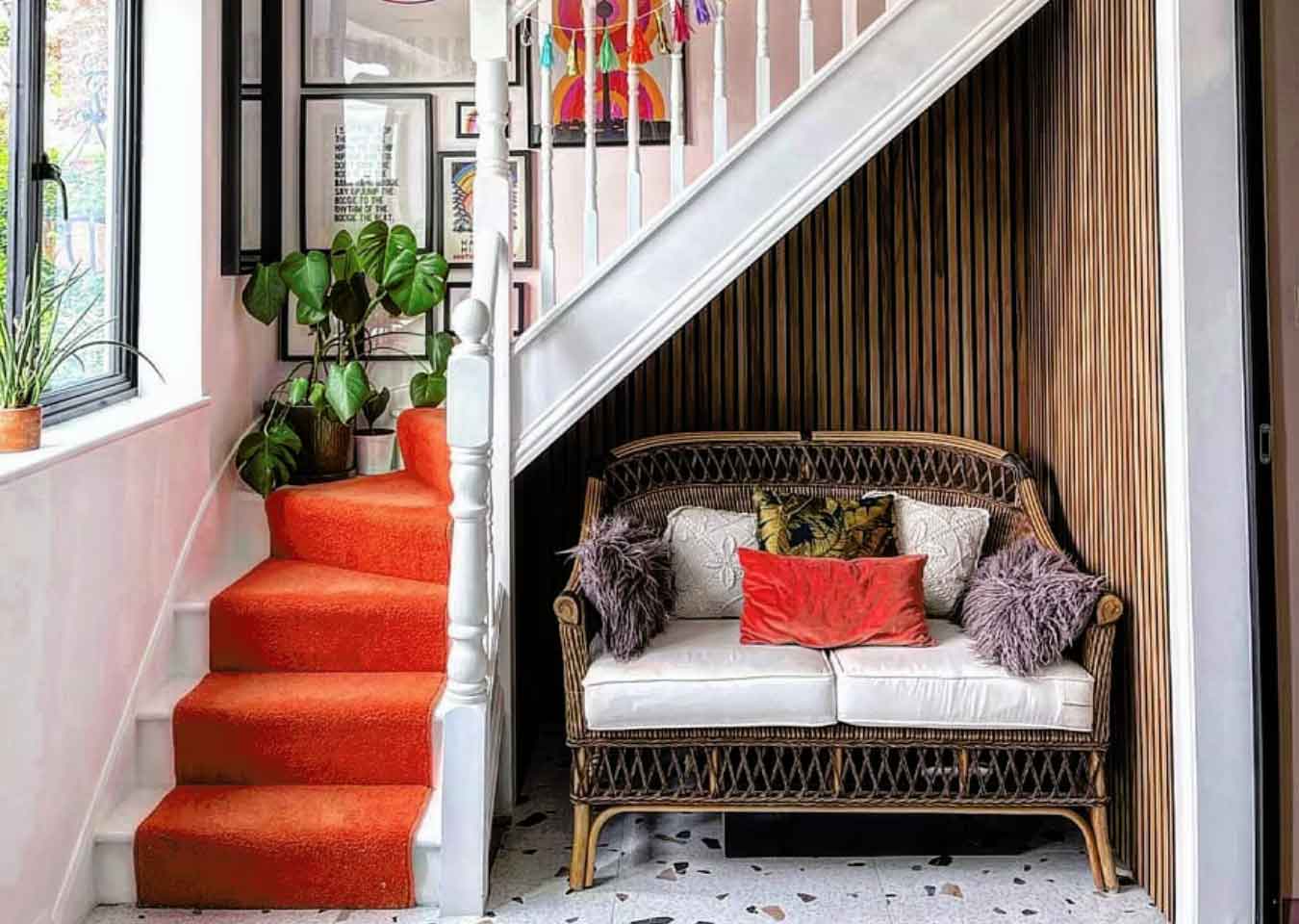 A white staircase with an orange runner, multicoloured wall art, green plants, wood panelling under the stairs and a rattan sofa.