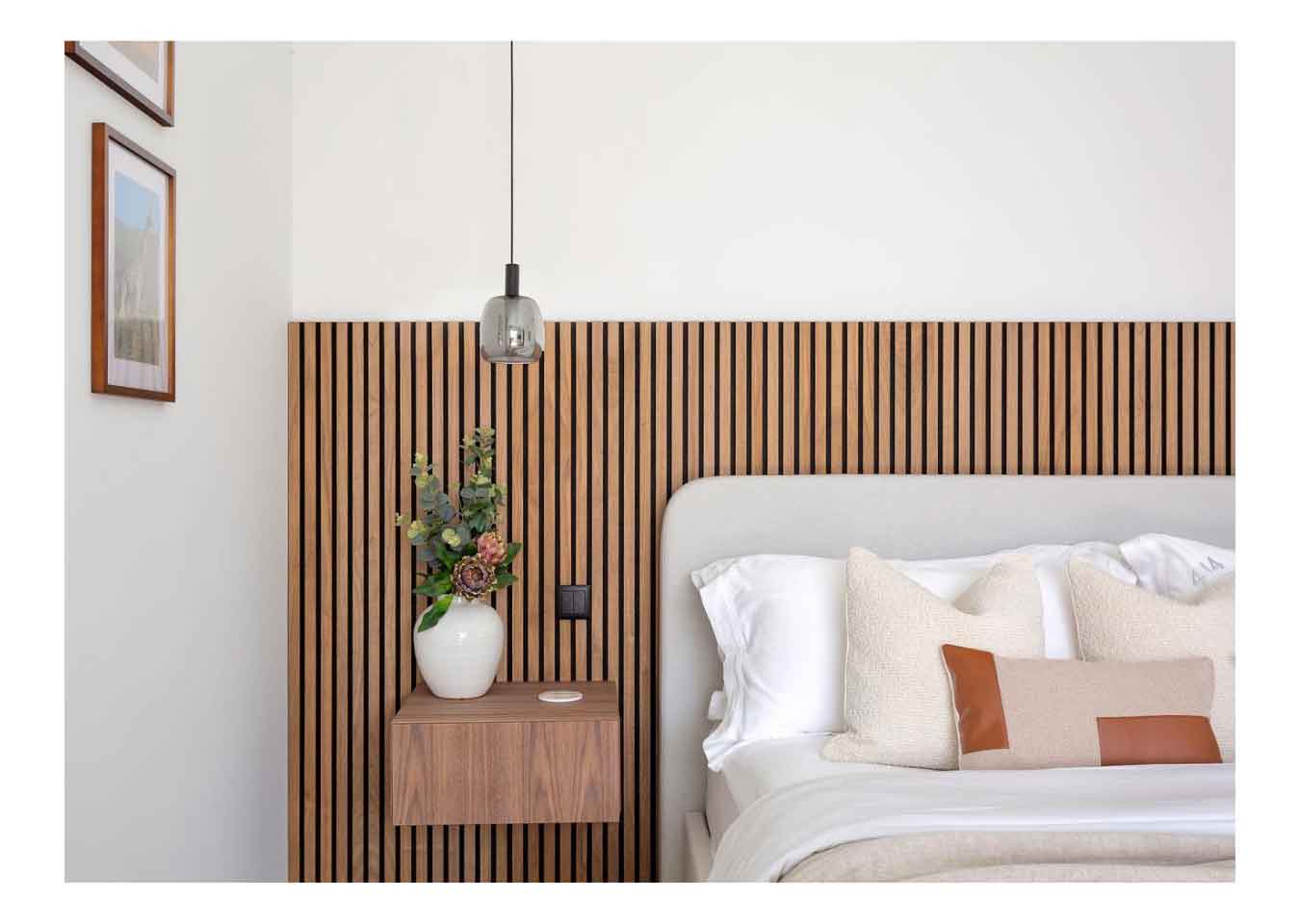 Bedroom with oak wood panelling, a grey and white bed, cream cushions, grey pendant light, a floating bedside table and vase of flowers.