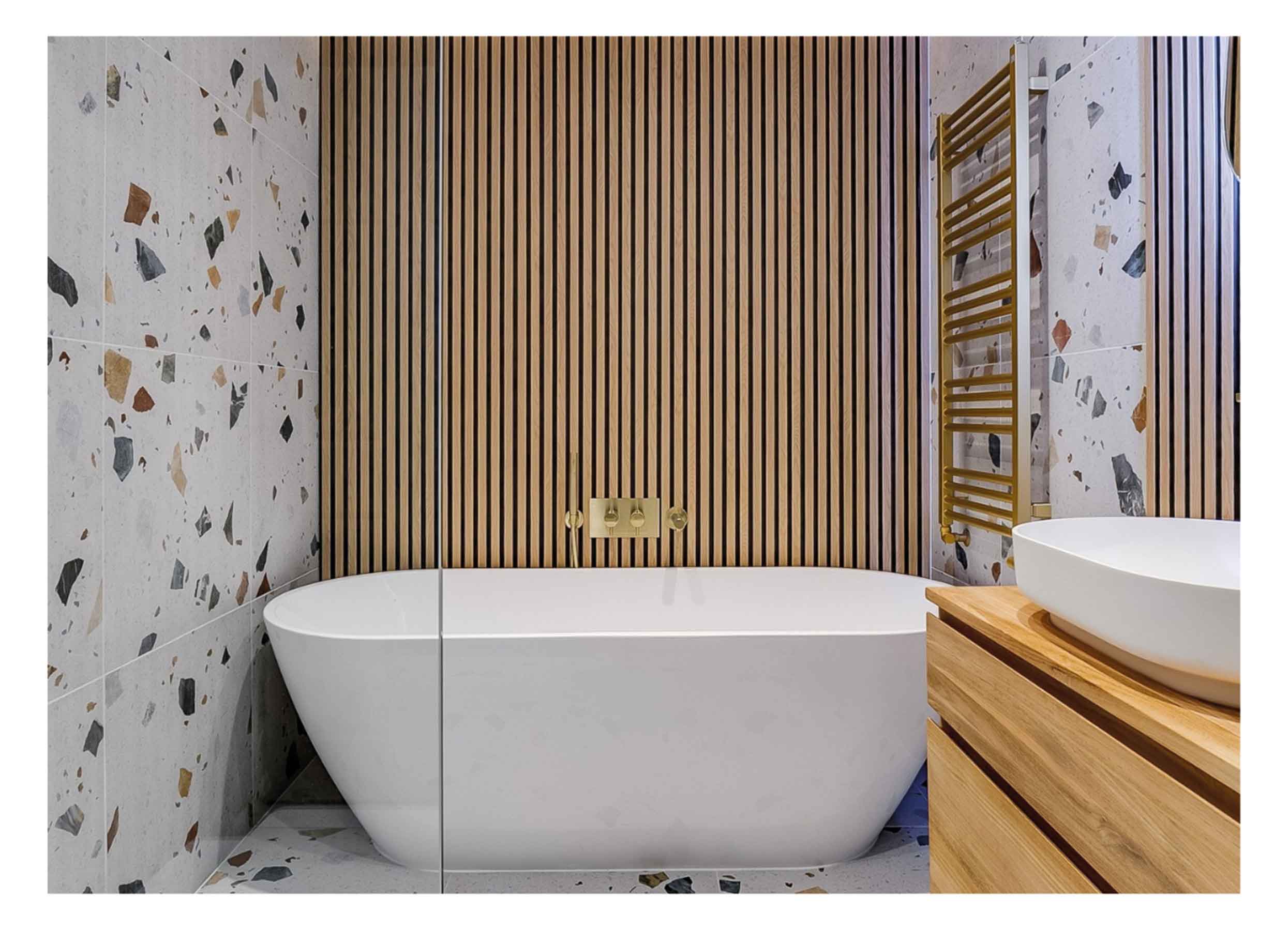 Brown wood panels and white terrazzo tiles on bathroom walls next to a white bathtub, gold-tone towel rail and sink unit.