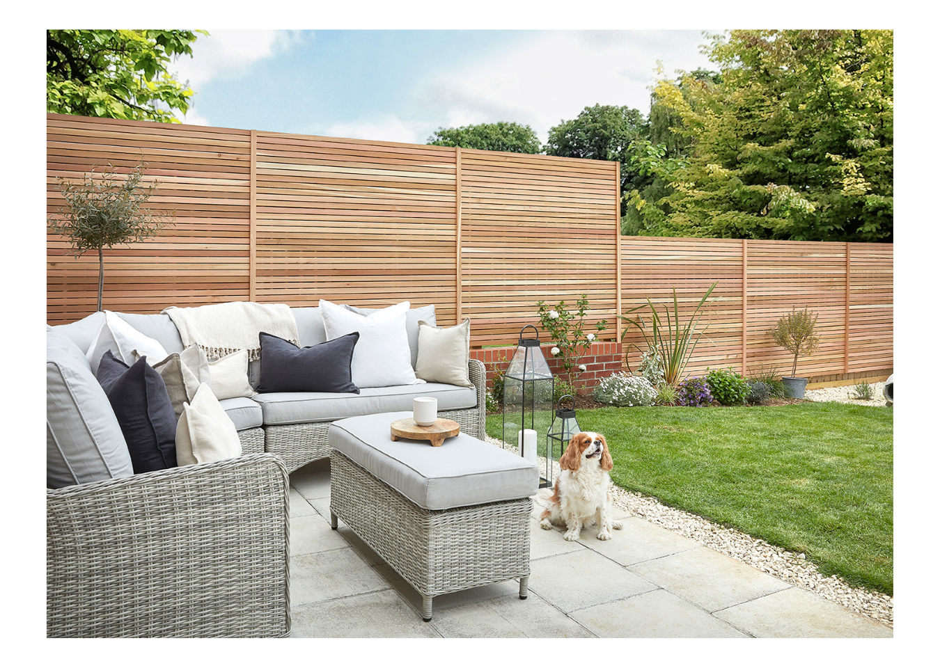 Outdoor entertaining area in a garden featuring a grey corner sofa and table with a dog sitting next to it.