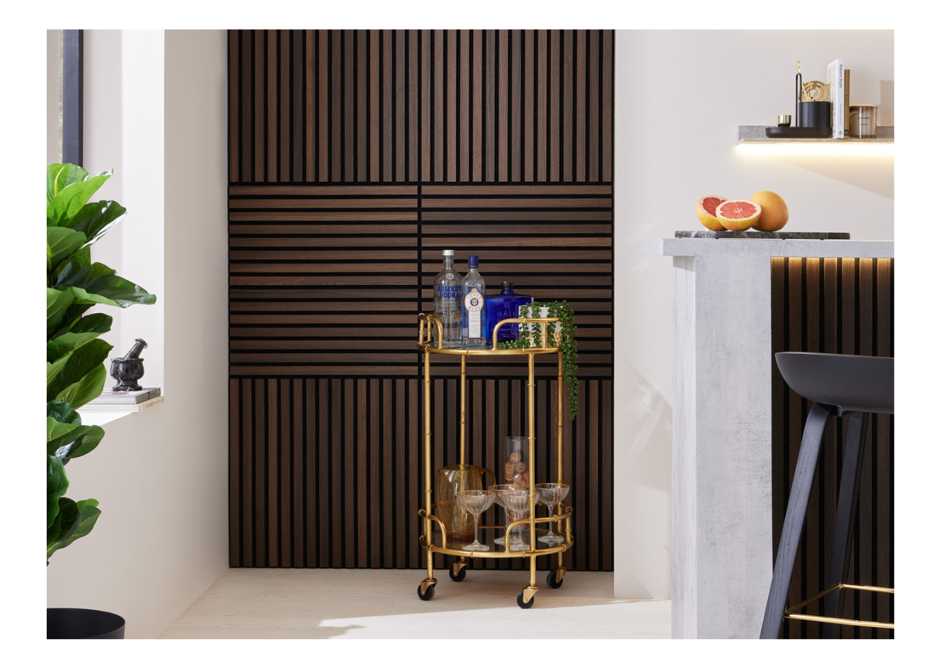 A gold-tone drinks trolley with bottles and glasses in front of SlatWall Mini Smoked Oak panels in a home bar.