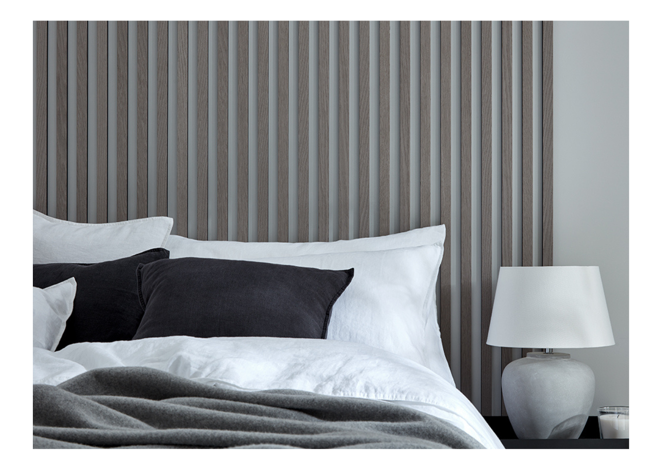 SlatWall Grey Oak wooden wall slats behind a white bed with black cushions and a grey blanket, next to a grey and white lamp.