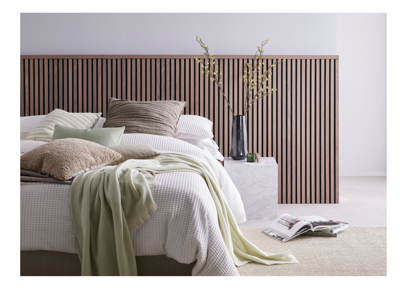 A white bed with beige cushions and a green blanket in front of half-wall panels made with SlatWall Midi Walnut and Top Trim.