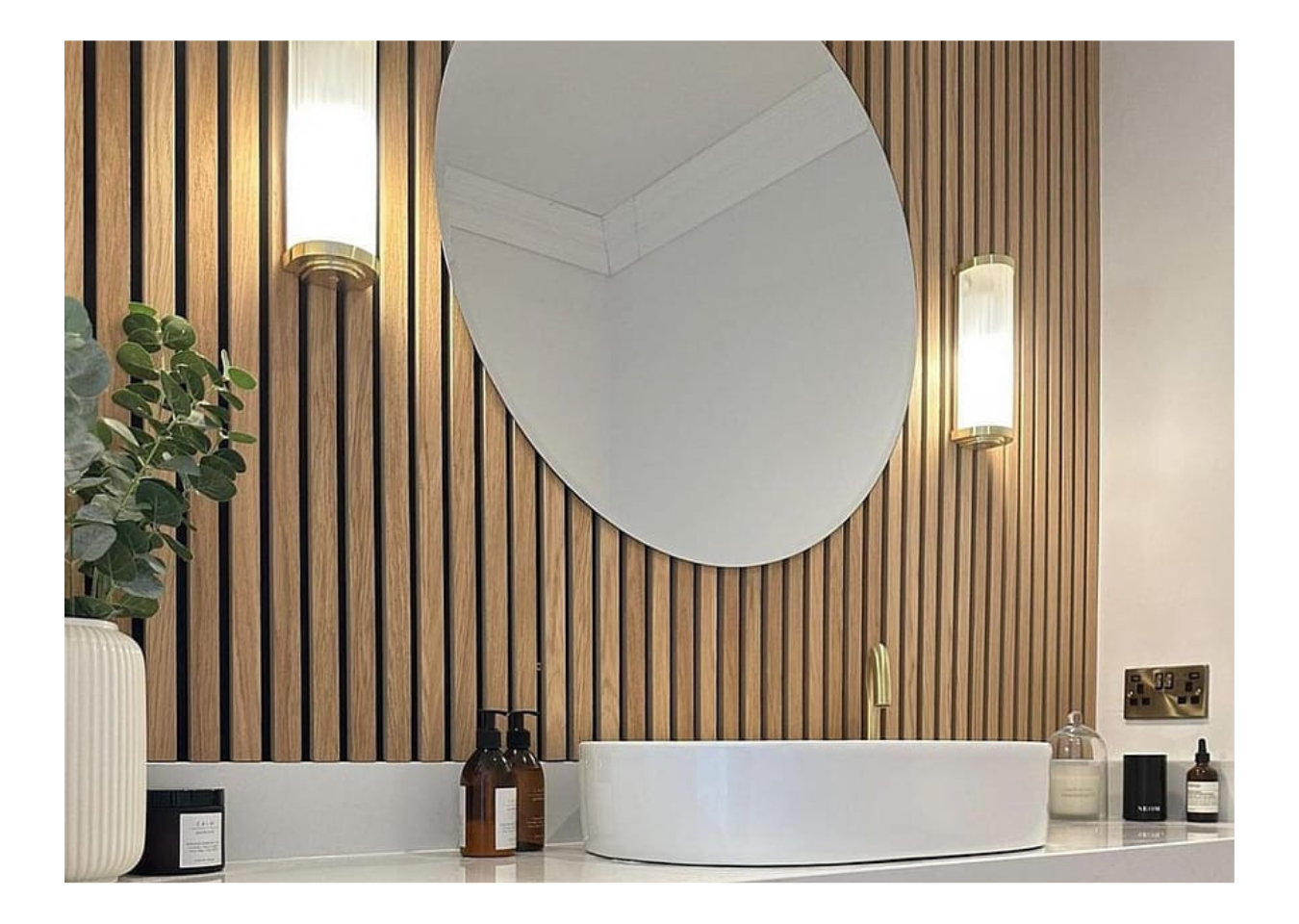 A large round mirror and two lights on a wall featuring SlatWall Waterproof Natural Oak and Black, above a white sink.