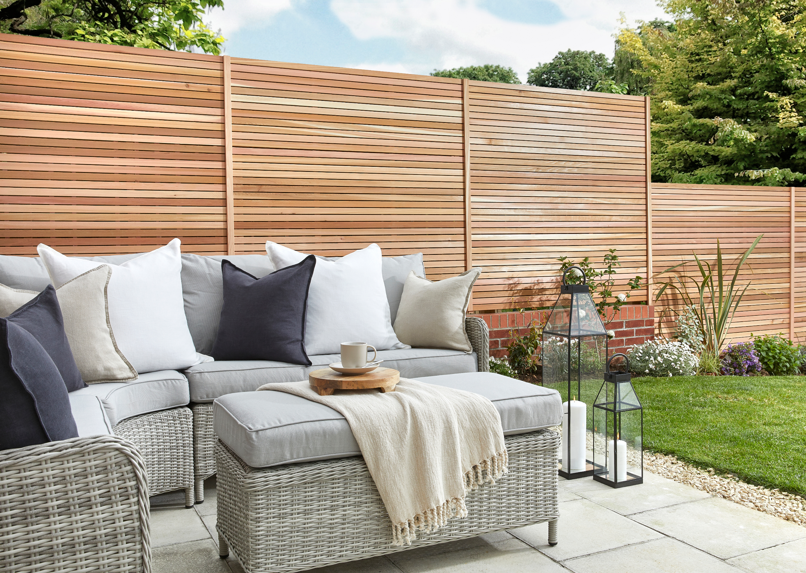 Outdoor seating area. Grey rattan sofas with white and grey soft furnishings. Cedar fence panels in the background