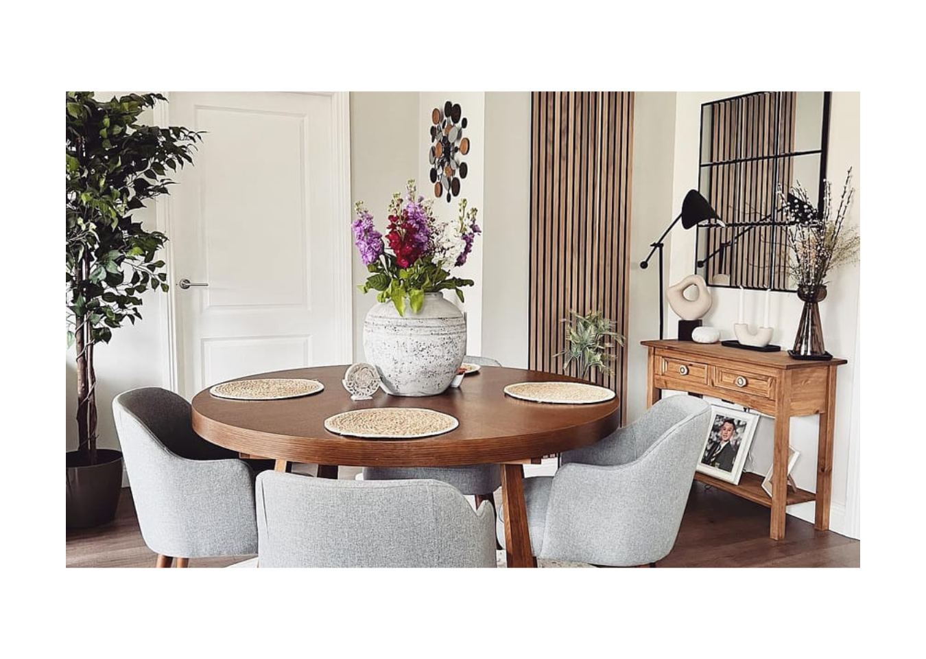 Single slatwall panel on wall besides a mirror. Round dining table in the foreground with grey chairs around and a floral centrepiece.