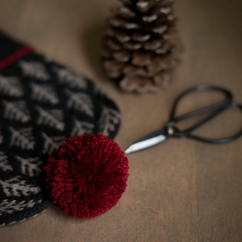 knit hat with red pom pom on a table with scissors and a pinecone