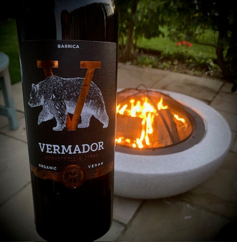 Photo shows a bottle of the wine with the warmth of a firepit in a lovely garden scene.