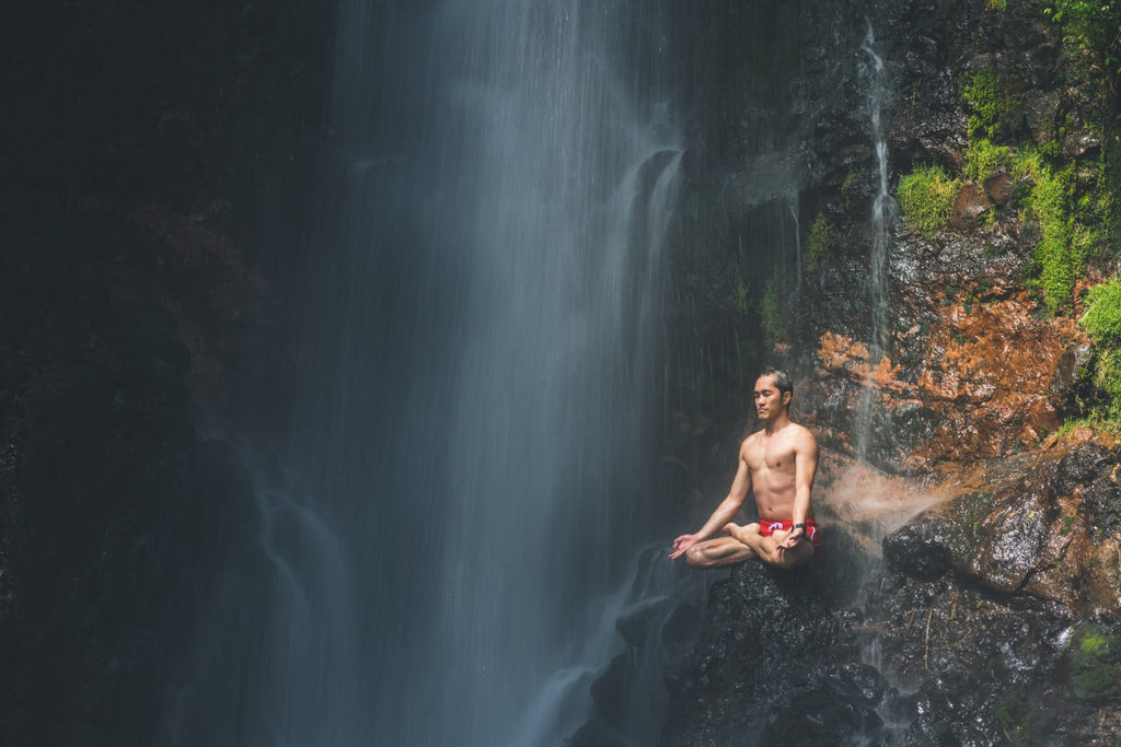 Young Man Meditating On Rocks Under The Flow Of Water Below Waterfall