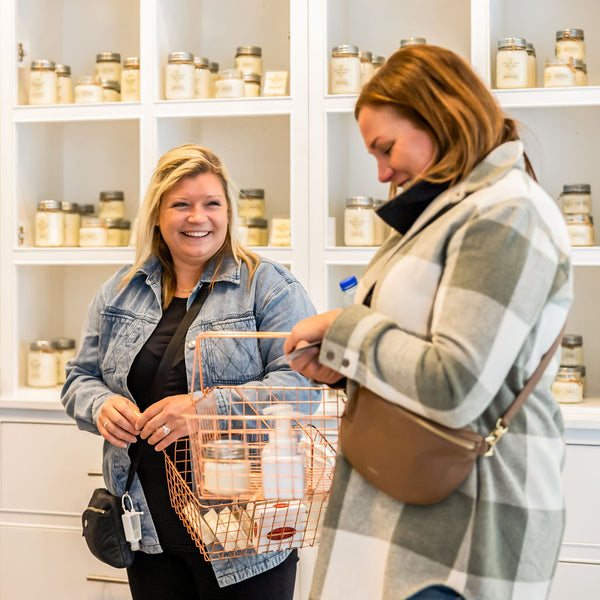 Two ladies stand inside the new Scented Market store in the Blue Mountain Village. They hold shopping baskets filled with products and are smiling.