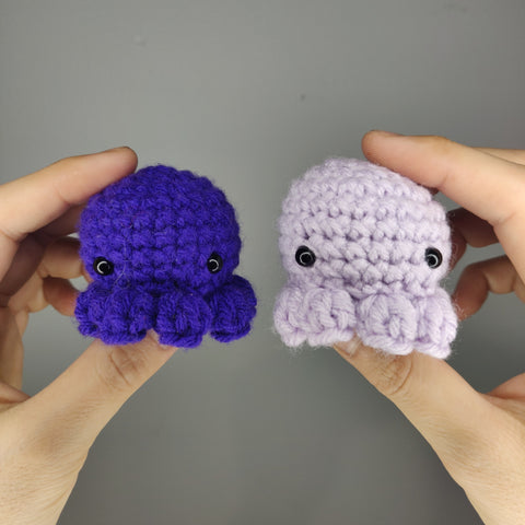 Pair of hands holding amigurumi octopus. The left one is a dark blue-purple, the right is lavender.