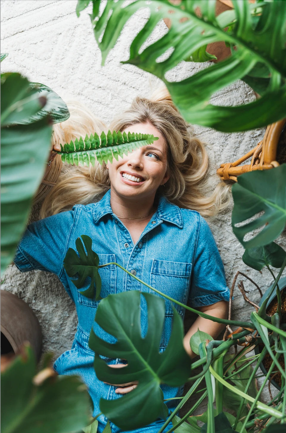 A woman with blond hair and a cheerful expression lies on her back among a variety of green houseplants, holding a fern leaf up to her eye like a playful monocle.