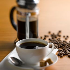 a mug of black coffee, in the background are a full french press and whole coffee beans to show what coffee for french press