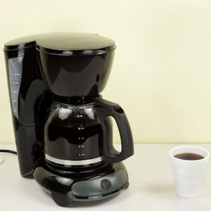 an electric drip coffee maker full of coffee, next to a styrofoam cup of black coffee