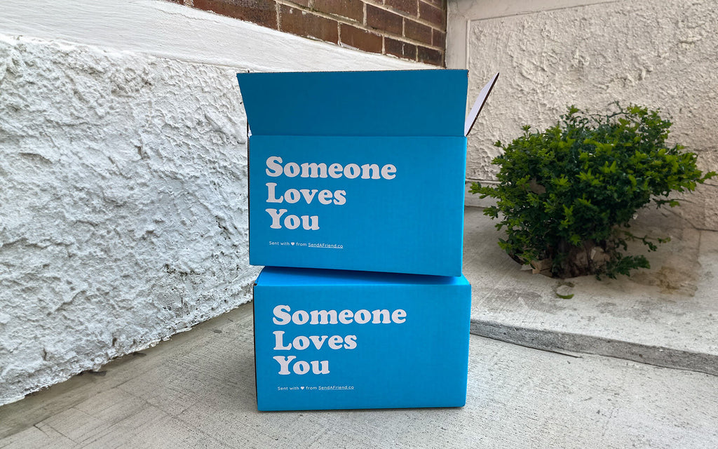 An image of two "Someone Loves You" boxes stacked sitting outdoors on concrete, one open, one not.