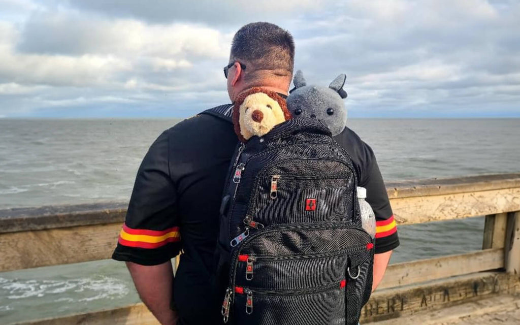 Pictured is a man standing on a boardwalk looking out at an ocean wearing a backpack with Oliver the Otter and Binks the Bat peeking their heads out of the open zipper