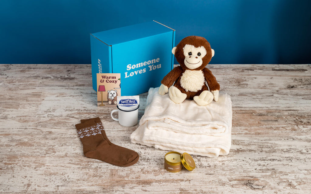 An image of SendAFriend's Cozy Bundle with Maria the Monkey: comfy blanket, a delightful candle, a pair of socks, mug with hot cocoa, and a "Warm & Cozy" card