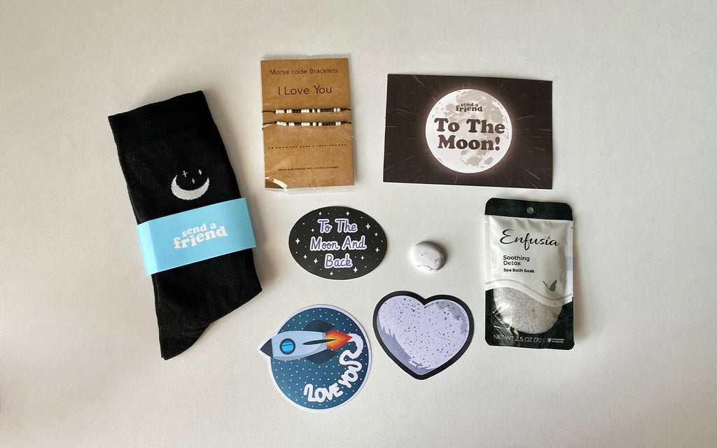 Pictured are items from the To the Moon and Back Bundle: moon socks, Morse Code bracelets, bath soak, a worry stone, and cosmic sticker pack