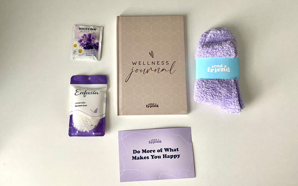 Pictured are items from the Self Care Bundle: fuzzy socks, a wellness journal, bath soak, a chamomile lavender tea bag, and curated Spotify playlist